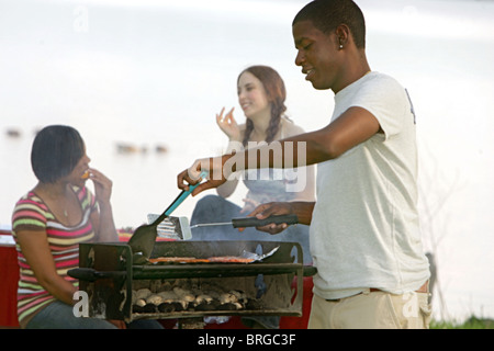 einem glücklichen zwanziger Jahre afrikanische amerikanische Mann Kochen auf ein Grill im Freien in einem Park mit zwei Freunden hinter Stockfoto