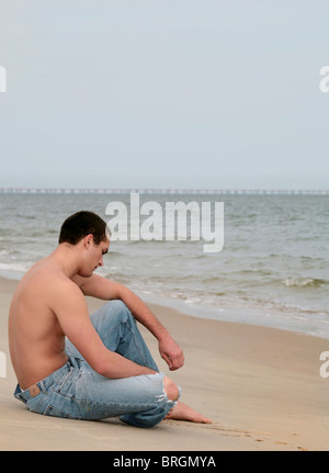 eine nachdenkliche traurig aussehende junge passen Mann sitzt auf einem Strand nahe dem Ozean Stockfoto