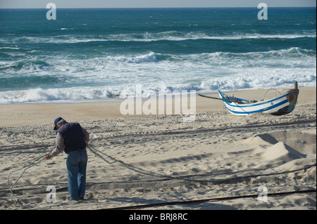 Fischer flickt die Netze mit einem traditionellen hölzernen Fischerboot und den Atlantischen Ozean als Hintergrund in Praia de Mira, Portugal Stockfoto