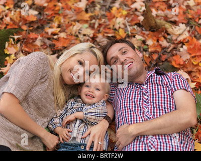 Glücklich lächelnde junge Eltern und ein zwei Jahre altes Mädchen auf bunten umgestürzten Baum liegen in der herbstlichen Natur lässt Stockfoto