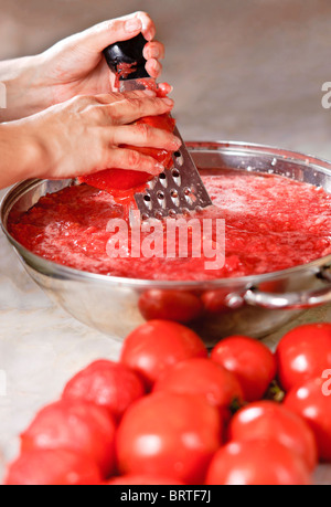 Eine Frauenhand reiben eine Tomate in großen, verchromten Topf. Tomaten stehen im Vordergrund Stockfoto