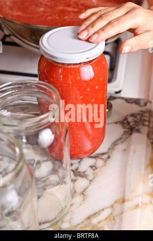 Die Hand einer Frau prüft die Vakuumdichtung eines Deckels auf Glas, gefüllt mit Tomatensaft. Ein leeres Glas ist in der Nähe auf einer Arbeitsplatte Küche Stockfoto