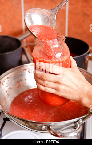 Die Hand einer Frau hält ein halb volles Glasglas während beheizten Tomatensaft aus einer Pfanne, über einem Topf gießen. Stockfoto