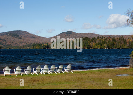 Acht weiße Adirondack Stühle in einer Reihe am Strand von Indian Lake. Blauer Himmel und Blick auf die Berge. Stockfoto