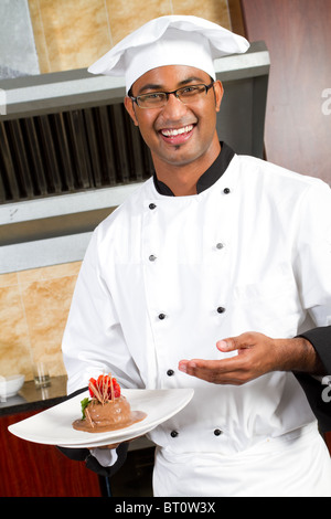 Junge glücklich männlicher indischer Koch präsentiert Dessert in Großküchen Stockfoto