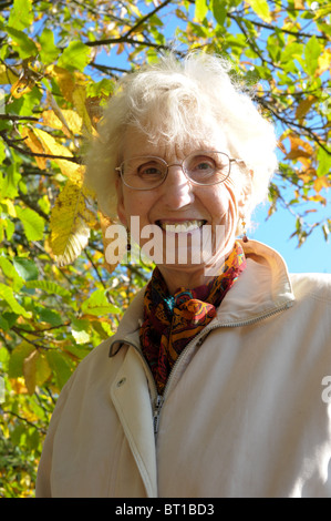 Weißhaarige ältere Dame lächelt in die Kamera mit goldenem Herbstlaub und blauer Himmel hinter ihr. Stockfoto
