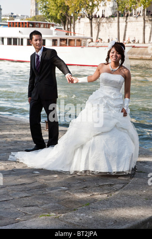 lächelnde Sonne Schecken asiatische Braut im trägerlosen Kleid & hübscher Bräutigam Pose für Portrait auf Paris Quai gegen blaue Wasser der Seine Stockfoto