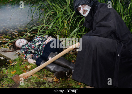 Mörderischen bösen Mann im schwarzen Mantel und weißes Gesicht mit Axt über einen toten Mädchen Körper von einem Teich hocken Stockfoto