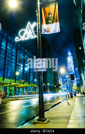 Broad Street at Night Philadelphia // PHILADELPHIA, Pennsylvania – die Broad Street wird nachts nach einem Regenschauer beleuchtet, während der nasse Bürgersteig die pulsierenden Lichter der Stadt reflektiert. Das Rathaus von Philadelphia ist im Hintergrund zu sehen, und Lichtspuren von vorbeifahrenden Fahrzeugen verleihen der Szene ein dynamisches Element. Stockfoto
