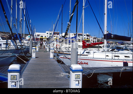 Der Club Mykonos Pier mit einer Vielzahl von teuren Yachten vor Anker und zwei Matrosen zu Fuß entlang der Pier in der Ferne. Stockfoto