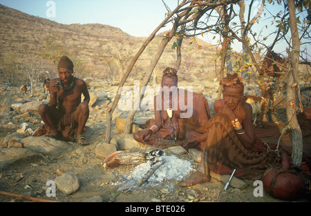 Himba-Mann und zwei ältere Frauen sitzen in der Nähe von der Asche des Feuers Stockfoto