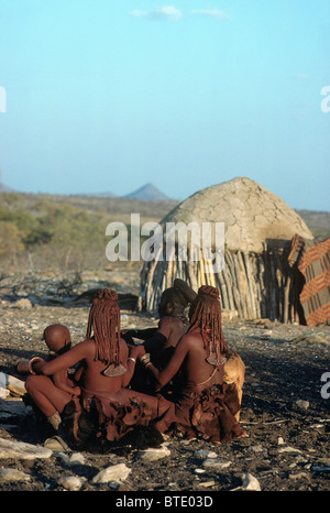 Drei Himba-Frauen und jungen Kleinkind sitzen auf dem Boden in der Nähe einer urige Hütte Stockfoto
