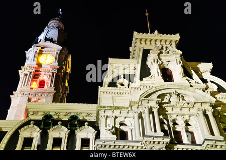 Außenansicht des Rathauses Philadelphia // PHILADELPHIA, Pennsylvania – ein Außendetail des Rathauses in Philadelphia, PA, bei Nacht. Der ikonische Uhrenturm ist hell beleuchtet und zeigt seine komplexen architektonischen Details und das sichtbare Zifferblatt. Die kunstvolle Fassade des Gebäudes, die mit Statuen und Schnitzereien verziert ist, hebt sich vom dunklen Himmel ab. Ein markantes rotes Licht verleiht dem unteren Teil des Turms einen lebendigen Touch. Stockfoto