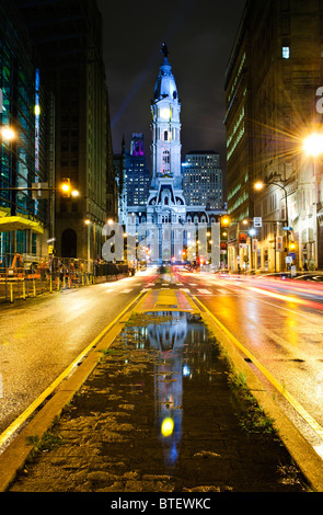Philadelphia City Hall Reflection Broad Street Philadelphia // PHILADELPHIA, Pennsylvania – das Philadelphia City Hall wird nachts von der Broad Street aus gesehen, nach Regen, mit lebhaften Verkehrsstreifen. Seine Reflexion ist deutlich sichtbar in einer Pfütze in der Mitte des Rahmens. Stockfoto