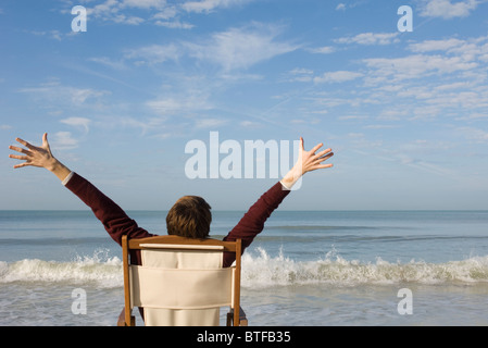 Junger Mann sitzt im Stuhl am Strand mit Arme angehoben, hintere Ansicht Stockfoto