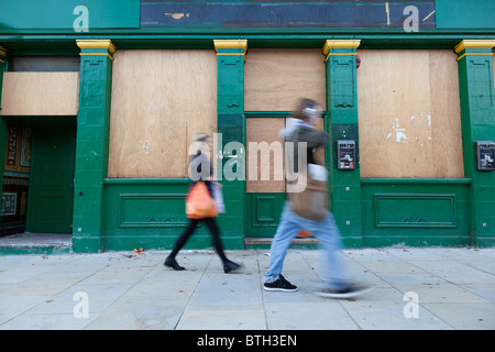 Ein Mann und eine Frau gehen vorbei an einem vernagelten Gebäude in London, England. Stockfoto