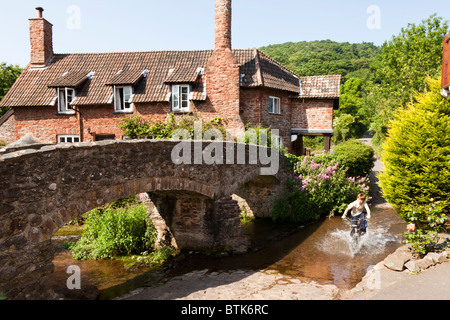 Eine junge Radsportler, die durch den Fluss Aller fließt unter der alten Pack Pferd Brücke bei Allerford, Exmoor, Somerset Stockfoto