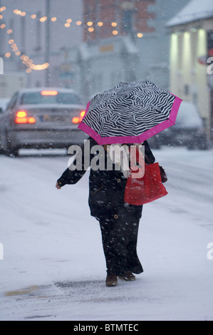 Eine Frau mit einem Regenschirm überquert eine Straße während einer Schneedusche in Castlebar, Co. Mayo, Irland Stockfoto