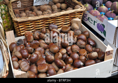 Eine Box stapelten sich mit Kastanien bereit zum Braten auf offenem Feuer, eine Schachtel mit Luxus-Feigen und einen Korb mit Walnüssen auf einem Marktstand Stockfoto