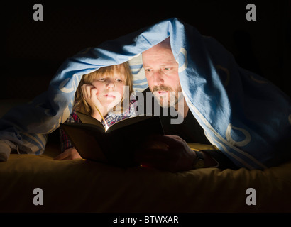 Vater liest eine Geschichte an seinen Sohn vor dem Schlafengehen mit Taschenlampe unter Bettwäsche. Stockfoto