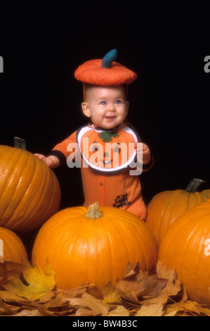 Acht Monate altes Baby Mädchen in Kürbis-Outfit Stand mit Kürbisse mit Herbstlaub im Innenbereich Stockfoto