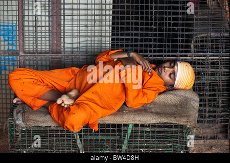 Indischer Junge in orange gekleidet schlafend auf einem Sack vor Metall Käfige für Haltungssysteme in verwendet werden. Die Metzger boy!. Andhra Pradesh, Indien Stockfoto