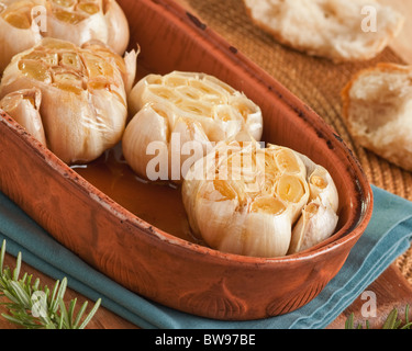 Frischer Knoblauch in Olivenöl in einen Tontopf mit knusprigen Baguette und Zweige Rosmarin sichtbar rösten Stockfoto