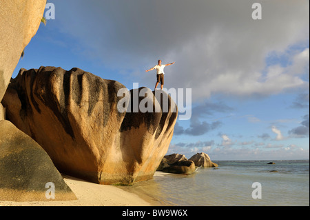 Mann mit Arme nach oben in der Luft stehend auf Granitfelsen auf Pointe Source D´ Argent, La Digue, Seychellen Stockfoto