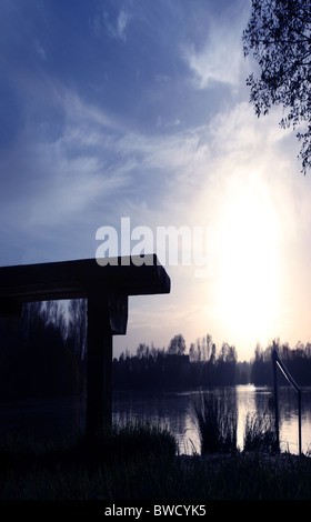 Leere Bank mit Blick auf einen See mit der Sonne einstellen und auf dem Wasser reflektiert Stockfoto