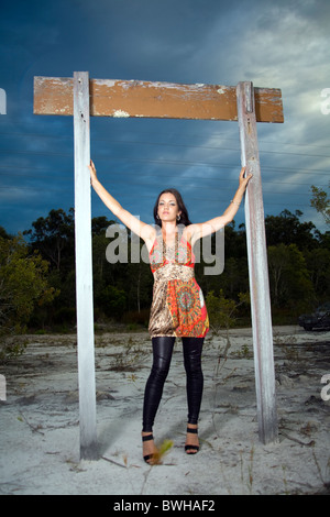 Attraktive junge Frau trägt eine helles Multi Farbe Minikleid mit schwarzen Strumpfhosen steht unter einer alten Holz Schild mit dunklen Wolke Stockfoto