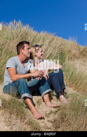 Ein junger Mann und Frau paar romantische zusammensitzen in grasbewachsenen Sanddünen von einem sonnigen Strand mit einem strahlend blauen Himmel Stockfoto