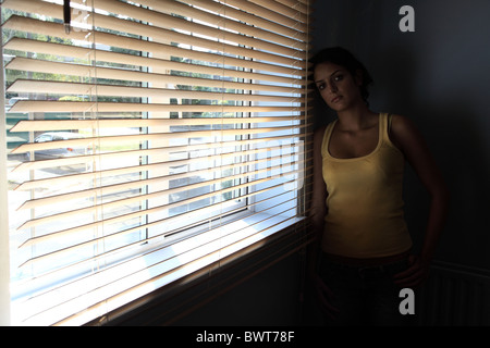 Junge Frau allein in einem dunklen Raum mit einer Jalousie, Blick in Richtung Kamera. Stockfoto