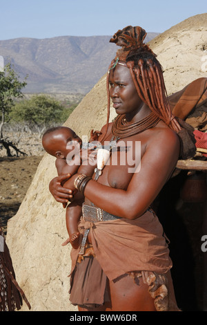 Himba-Frau mit "Oruvanda" traditionelle Leder Kopf Ornament und Ocker überdachte Flechten Frisur, halten Baby außerhalb Lehmhütte, Stockfoto