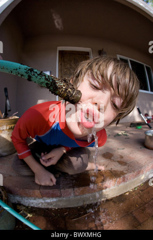 Acht Jahre alter Junge Trinkwasser aus einem Schlauch in Schlamm bedeckt. Stockfoto