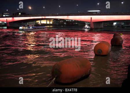 Boote passieren nachts unter London Brücke, die am Abend rot leuchtet. Dies ist eines der am häufigsten verwendeten Überfahrten des Flusses Themse. Stockfoto