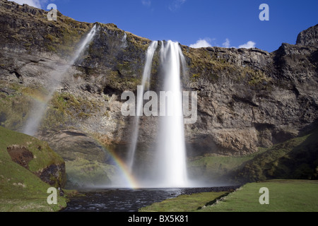 Seljalandsfoss Wasserfall über hoch aufragenden Klippen in hellem Sonnenlicht mit Regenbogen am Fuße des Wasserfalls, Island Stockfoto