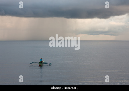 Ein Mann sitzt friedlich in einem traditionellen philippinischen Fischerboot mit einer Regendusche in der Ferne. Stockfoto