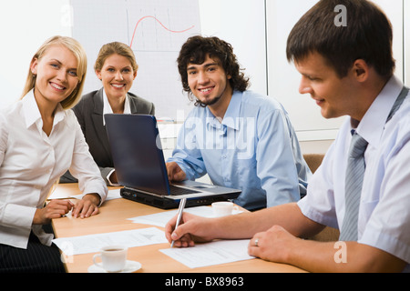 Porträt von vier junge Geschäftsleute sitzen um den Tisch mit Dokumenten, Papieren, Kugelschreiber, Tasse Kaffee und am Laptop eröffnet Stockfoto