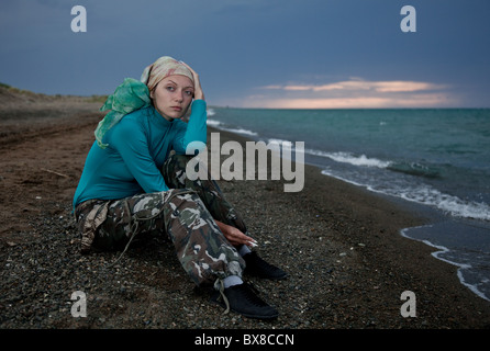 Das schöne Mädchen sitzt am Ufer des Meeres auf einem absteigenden Ast, hält eine Zigarette in der Hand und sieht irgendwo Stockfoto