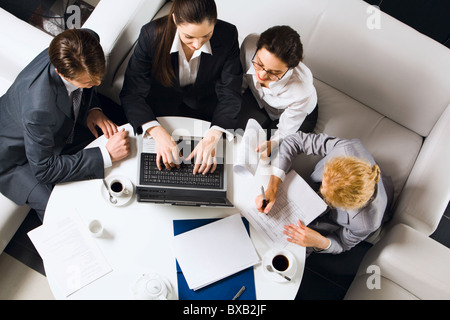 Drei Unternehmerinnen und Unternehmer sitzen auf den weißen Sofas am runden Tisch mit geöffneten Laptop, Dokumente und Tassen von co Stockfoto