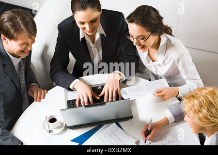 Drei lachende Unternehmerinnen und Unternehmer sitzen auf den weißen Sofas am runden Tisch mit geöffneten Laptop, Dokumente und c Stockfoto