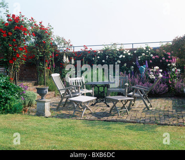 Holztisch mit Stühlen auf Backstein gepflasterte Terrasse im ummauerten Garten mit roten Kletterrosen auf Bogen und weißen Rosen wachsen auf Wand Stockfoto