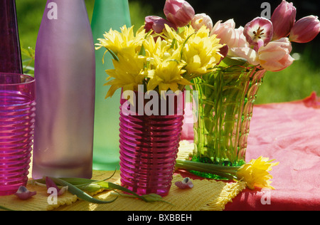 Stillleben mit gelben Doppelte Narzissen und rosa Tulpen in hell rosa und grün Vasen neben lila und grüne Glasflaschen Stockfoto