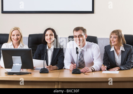 Erfolgreiche lächelnde Menschen sitzen in einer Reihe in schwarze bequeme Stühle am Tisch mit Monitor, Mikrofone und Dokumenten auf i Stockfoto