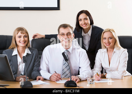 Drei erfolgreiche lächelnde Menschen sitzen in einer Reihe in schwarze bequeme Stühle am Tisch mit Monitor, Mikrofonen und Dokument Stockfoto