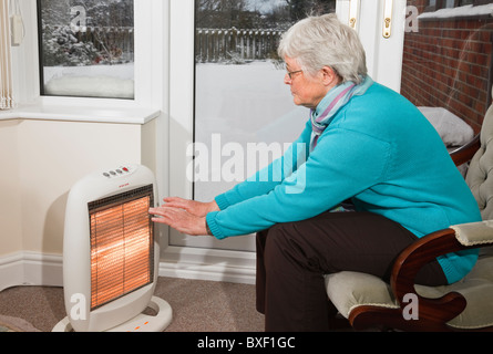 Eine ältere Frau Rentner OAP versuchen, warmen wärmenden Händen drinnen saß vor einer Halogen elektrische Feuer mit Schnee draußen ein Fenster im Winter UK zu halten Stockfoto