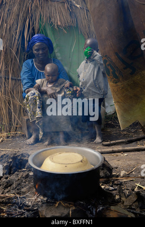 Eine intern vertriebene Frau mit ihren Kindern heizt vor einer provisorischen Strohhütte in einem IDP-Lager in der Provinz Nord-Kivu, DR Kongo, Afrika, Nahrung über Lagerfeuer auf Stockfoto