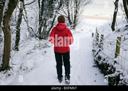 Einsame Frau Walker in roter Jacke gehen auf Schnee bedeckt die Insel Anglesey Coastal Weg zum Strand im Winter. North Wales, UK. Stockfoto