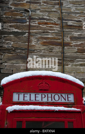 Dach und oberen Abschnitt von einer traditionellen BT-rote Telefonzelle, mit Schnee auf der Oberseite und einer Steinmauer hinter sich Stockfoto