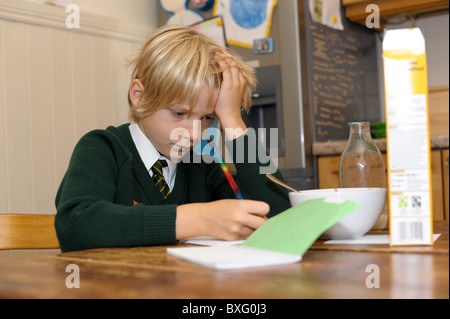 Ein kleiner Junge seine Schuluniform tragen kämpft mit seinen Hausaufgaben sitzen am Küchentisch schreibt in seinem Buch beim Frühstück Stockfoto